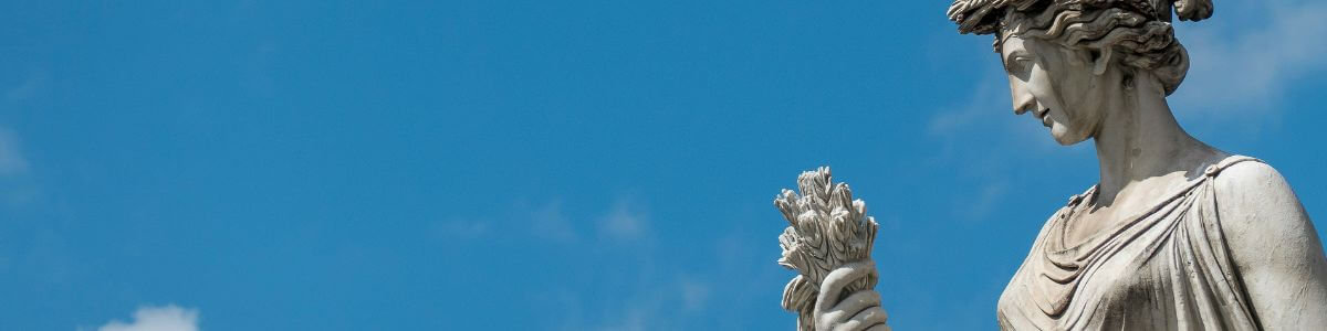 Ancient statue of a woman holding flowers, gazing at them against a blue sky.